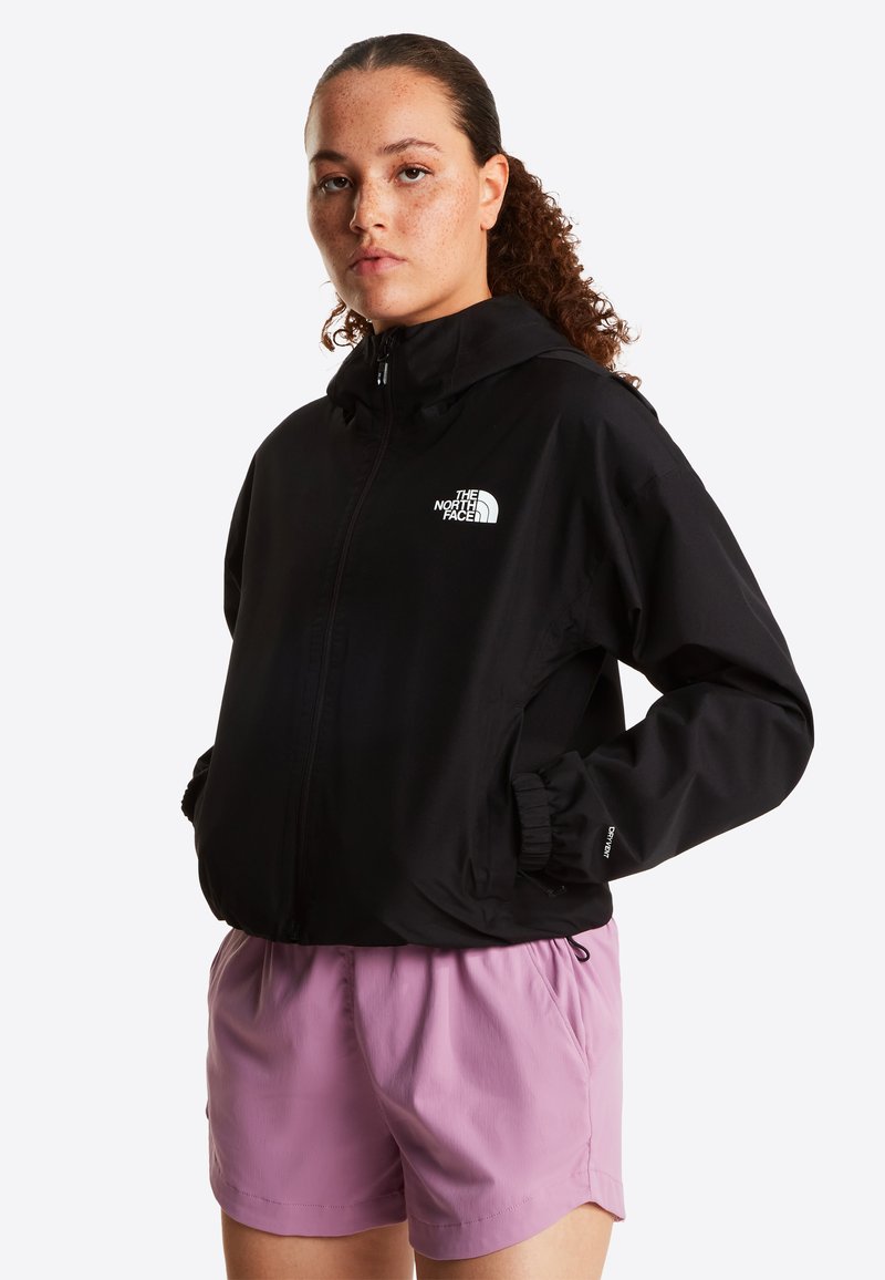 Νεαρή γυναίκα με κατσαρά μαλλιά που φοράει μαύρο μπουφάν North Face και ανοιχτόχρωμο μωβ σορτς, με τα χέρια στις τσέπες, ουδέτερη έκφραση, λευκό φόντο.