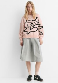 Maglione rosa lavorato a maglia con motivo astratto nero, abbinato a una gonna a pieghe grigia. Sneakers nere e calzini leggeri completano l'outfit.