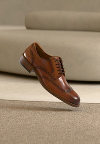 Καφέ δερμάτινο παπούτσι dress με φτερά, αιωρούμενο πάνω από μπεζ χαλί, με έναν ανοιχτόχρωμο καναπέ θολωμένο στο παρασκήνιο.