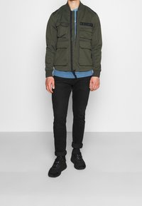 Olivgrön bomberjacka med flera framfickor, dragkedjestängning och ribbade ärmslut, bärs över en ljusblå skjorta, ihop med svarta jeans och sneakers.