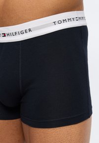 Κοντινό πλάνο ενός ατόμου που φοράει μαύρα μποξεράκια Tommy Hilfiger με λευκή μέση που φέρει το όνομα και το λογότυπο της μάρκας.