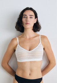 White bralette with a textured, wavy pattern; soft fabric; v-neck design; adjustable straps; worn with black underwear.