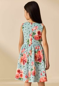 Fille aux longs cheveux foncés portant une robe sans manches bleu clair à pois noirs et motifs floraux roses, tournée de dos.