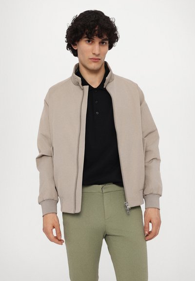 Beige lätt bomberjacka med ståkrage, ribbade manschetter och dragkedja, bärs över en svart polo och olivgröna byxor.