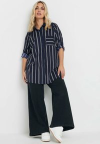 Camicia a righe navy con bottoni e maniche arrotolate, abbinata a pantaloni neri larghi e sneakers bianche. Presenta una tasca frontale e una vestibilità comoda.