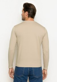 Beżowy t-shirt z długim rękawem, wykonany z miękkiego materiału, z okrągłym dekoltem i przeszytym wykończeniem. Noszony z niebieskimi dżinsami.