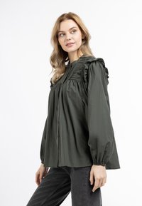 Donkergroene blouse met knopen aan de voorkant, ruches bij de schouders en een zachte textuur. Beschikt over geplooide details langs de halslijn en puffmouwen.