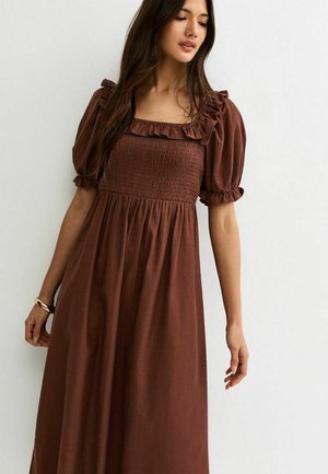 Femme portant une robe midi marron avec des manches bouffantes et un corsage smocké, debout sur un fond blanc uni.
