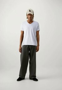 O tânăr îmbrăcat cu un tricou alb cu guler în V, pantaloni largi gri închis, pantofi negri și o șapcă de baseball bej cu inscripția "LA", stând cu fața înainte.