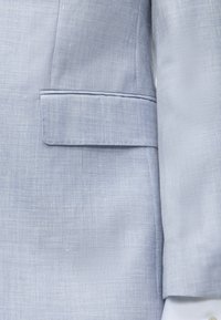 Gros plan d'une veste de costume en tissu bleu clair texturé avec une poche à rabat et une partie de la manche visible.