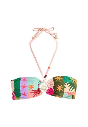 Osoba z krótkimi brązowymi włosami stoi na plaży, ubrana w kolorowy top bikini na ramiączkach z zawieszkami w kształcie słońca, związanymi na szyi.