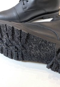 Zapato negro de cuero con suela de goma texturizada, que presenta un patrón de tracción resistente y un diseño minimalista; incluye cordones y detalles de costura.