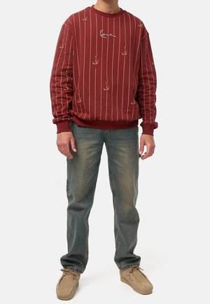 Mężczyzna w czerwonym swetrze z cienkimi pionowymi paskami i ręcznie rysowanymi twarzami, niebieskich dżinsach oraz jasnobrązowych zamszowych butach, stojący na białym tle.