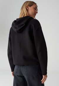 Felpa nera con cappuccio e vestibilità rilassata, realizzata in tessuto morbido. Dotata di un ampio cappuccio e polsini a costine. Abbinata a jeans denim scuro.