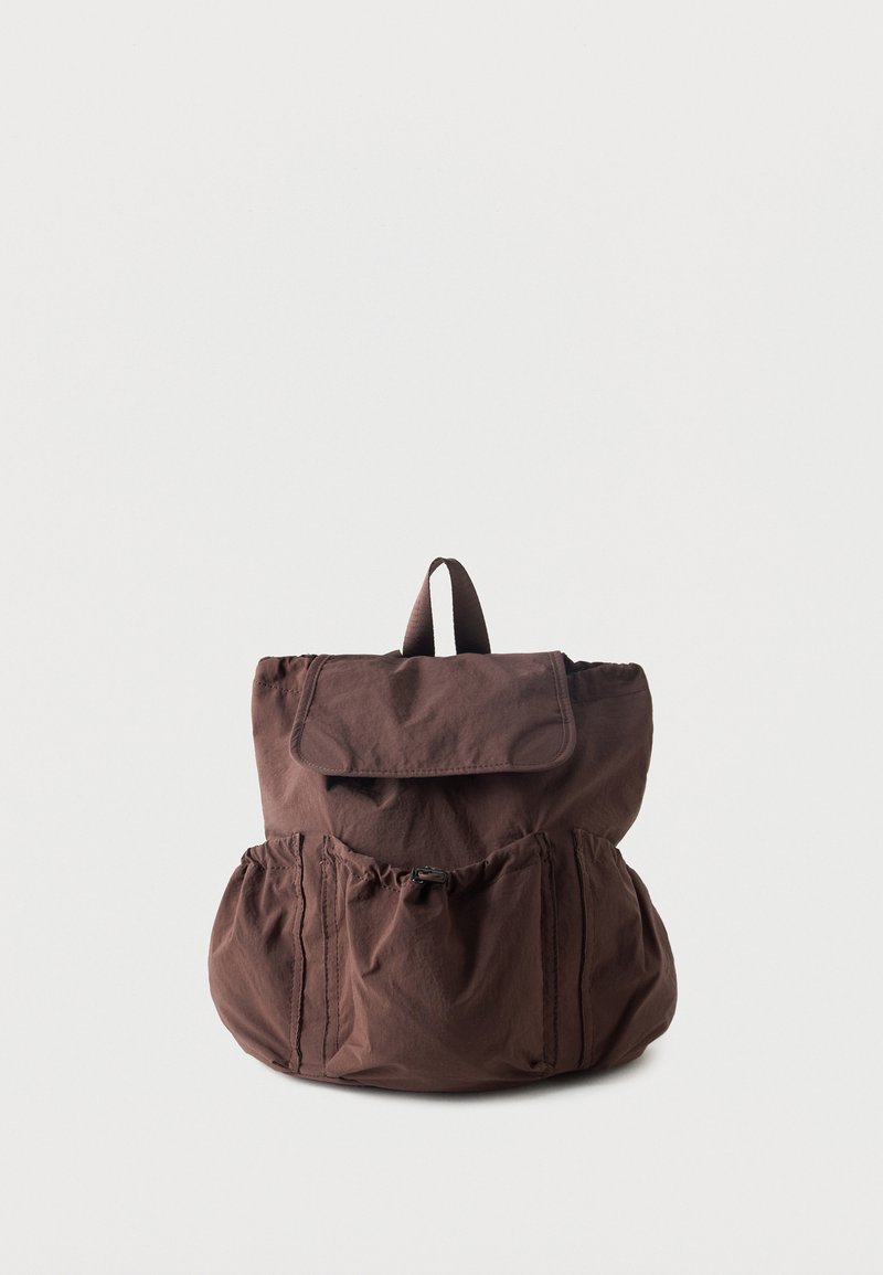 Καφέ υφασμάτινο σακίδιο με καπάκι στην κορυφή, μπροστινή τσέπη με κλείσιμο με πόρπη, δύο πλαϊνές τσέπες και ένα μικρό χερούλι στο πάνω μέρος, σε λευκό φόντο.