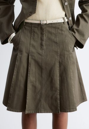 Femme portant une jupe plissée vert olive avec les mains dans les poches, un haut côtelé beige rentré, et une fine ceinture beige clair visible.