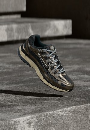 Tmavý kovový a černý běžecký bot Nike s béžovou podrážkou plovoucí nad šedými betonovými schody.