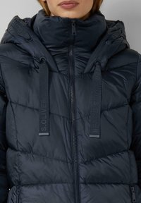 Doudoune bleu marine avec un col monté, deux poches avant zippées, des cordons ajustables et un motif matelassé texturé. Fermeture éclair.