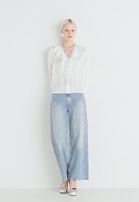 Blusa bianca con davanti arricciato, abbinata a jeans in denim azzurro a gamba larga. Modella indossa sandali con zeppa argento. Estetica semplice e minimalista.