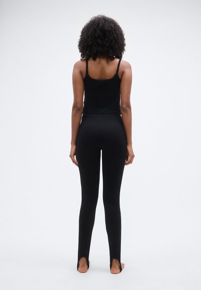 Débardeur noir à fines bretelles, associé à des leggings noirs moulants avec une fente en bas. Texture lisse partout.