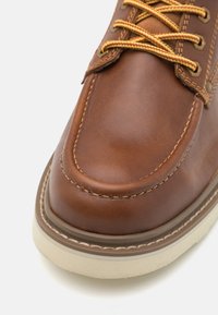 Bota de cuero marrón con punta redondeada, costuras en contraste en color crema y cordones amarillos. La suela es de goma de color claro, proporcionando una base resistente.