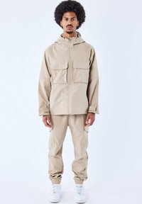 Beige jasje met twee voorzakken en bijpassende broek. De stof lijkt glad, met elastische manchetten aan de broek. Witte sneakers maken de look compleet.