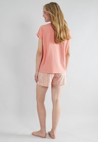 Koraalkleurig katoen T-shirt met opgerolde mouwen, gecombineerd met roze kortebroek met bloemenpatroon. Model staat blootsvoets, iets van de camera af gedraaid.