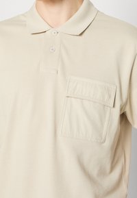 Polo shirt beige en tissu texturé, avec un col replié, trois boutons et une poche à patch sur la poitrine gauche.