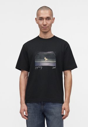 Homme à la tête rasée portant un t-shirt noir avec un graphisme d'un petit animal dans un cadre rectangulaire et un jean bleu.