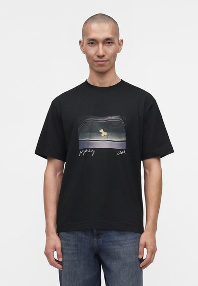 Homme à la tête rasée portant un t-shirt noir avec un graphisme d'un petit animal dans un cadre rectangulaire et un jean bleu.