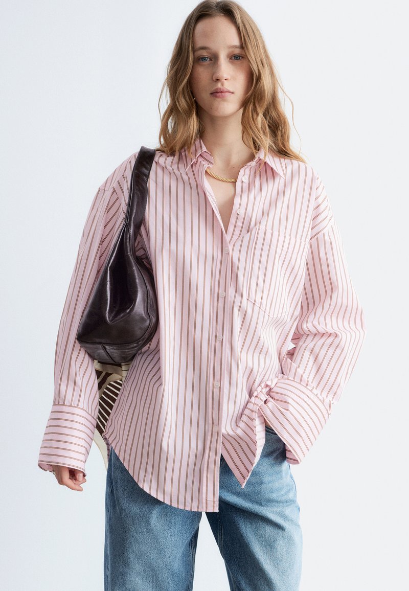 Jeune femme portant une chemise rose à rayures amples, un jean bleu, un collier en or et un sac à bandoulière noir, posant devant un fond uni.
