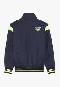 Námořnická modrá bunda s vysokým límcem, neonově žlutými akcenty na ramenou a pruhovanými žebrovanými manžetami. Na zadní straně je potisk "CARS JEANS 1982".