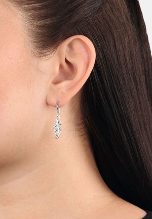 Primer plano de la oreja de una mujer con cabello oscuro que lleva un pendiente colgante en forma de hoja de plata.