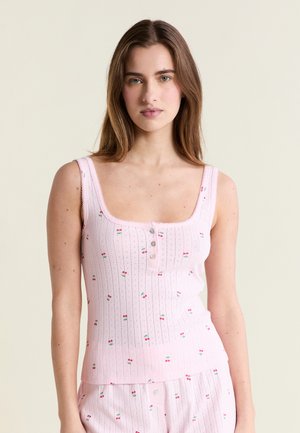 Jeune femme aux longs cheveux bruns portant un haut rose sans manches et un short assorti avec un petit imprimé cerises et des boutons.