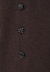 Tmavě hnědá látka s texturovaným povrchem, na přední straně se čtyřmi černými knoflíky. Knoflíky mají čtyřdírový design.