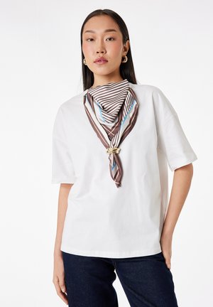 Vrouw met lang donker haar draagt een wit topje met korte mouwen, een gestreepte bruine en blauwe sjaal met gouden ring en donkerblauwe spijkerbroek tegen een witte achtergrond.