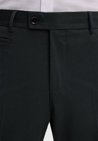Donkergroene broek van gladde stof, met een enkele voorplooien, riemlussen en een knoopsluiting. Inclusief een zijzak.