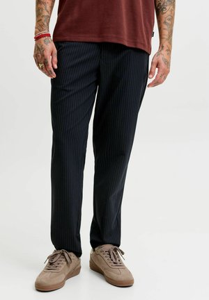 Homme portant un pantalon noir à fines rayures, des baskets marron et une chemise bordeaux, debout avec les bras tatoués visibles sur un fond uni.