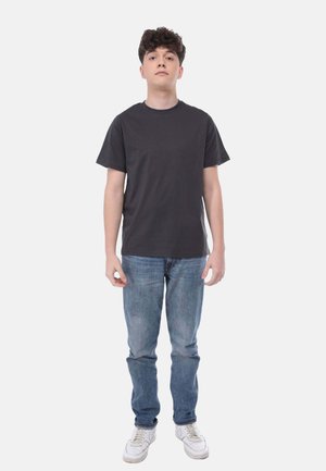 Schwarzes Baumwoll-T-Shirt, kurze Ärmel, Rundhalsausschnitt, kombiniert mit hellblauen Jeans. Das Model trägt weiße Sneaker. Der Hintergrund ist weiß.