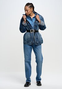 Jeansjacke mit tailliertem Design, ausgestattet mit einer Knopfleiste und mehreren Taschen, kombiniert mit blauen Jeans und schwarzen Schuhen.