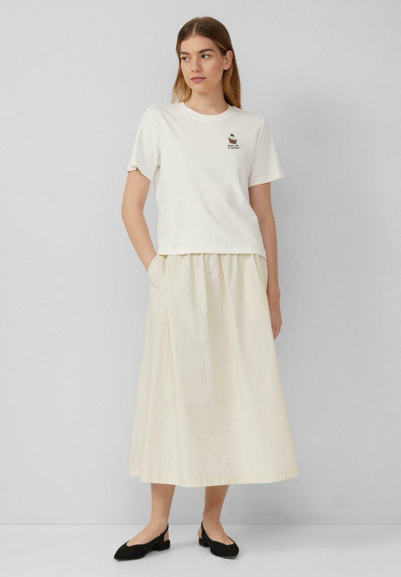 T-shirt en coton blanc à manches courtes avec un design brodé, associé à une jupe fluide beige clair longueur cheville. Chaussures noires.