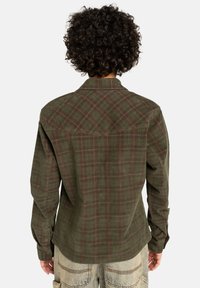Persona con capelli ricci che indossa una camicia a maniche lunghe a quadri verde e marrone e pantaloni in denim blu chiaro, vista da dietro.