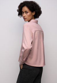Blusa rosa chiaro con colletto, maniche lunghe e schiena plissée. Il tessuto appare liscio con una vestibilità leggermente rilassata. Indossata con pantaloni scuri e testurizzati.