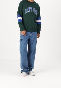 Sweatshirt vert avec l'impression "GANT 1949", présentant des rayures bleues et blanches sur les manches. Associé à un jean cargo bleu clair et des baskets blanches.
