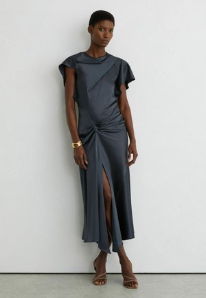 Žena v tmavosivej asymetrickej midišate s bočným rozparkom, detailom s volánikmi na rukáve, zlatým náramkom a otvorenými lodičkami stojaca pri bielej stene.