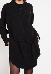 Vestido negro de camisa con botones, de manga larga, con dos bolsillos en el pecho y un dobladillo curvado. Presenta una cintura con cordón y un detalle de cremallera.