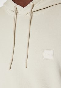 Krämfärgad huvtröja i mjukt tyg, med dragsko i huvan och en subtil fyrkantig logotyp med texten "BOSS."