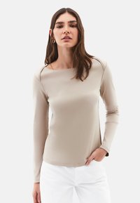 Frau mit langen braunen Haaren, die goldene Creolen, ein beige Langarmoberteil und weiße Hosen trägt, mit einer Hand in der Tasche.