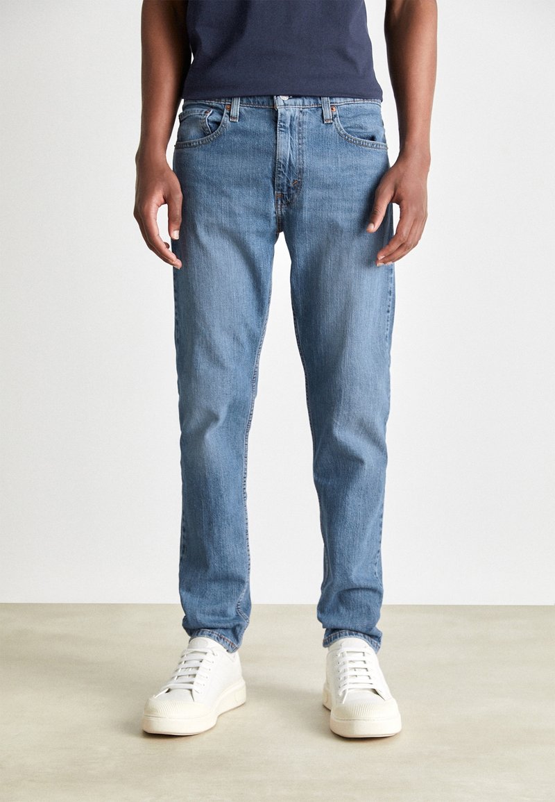 Homme portant un jean bleu et des baskets blanches debout sur un sol clair devant un fond blanc uni.
