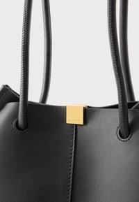 Borsa a mano in pelle nera con due manici cuciti e una piccola chiusura quadrata dorata incisa con il nome del marchio.